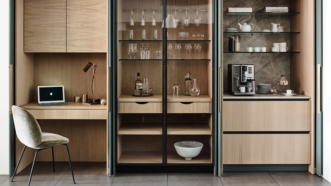 Comprehensive timber storage wall featuring oak cabinetry, glass-fronted display units with glassware and ceramics, integrated desk area with laptop, grey upholstered chair, and "For Your Home" messaging promoting bespoke furniture beyond kitchens with "VIEW GALLERY" call-to-action