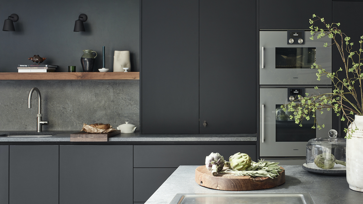 Matte charcoal kitchen with flat-front cabinets, grey stone worktop and splashback, walnut open shelf, stainless double ovens, and a sink; artichokes and branches on the island.