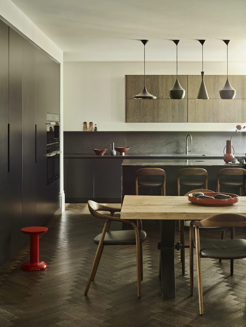 Open-plan dining kitchen with dark cabinetry, long stone worktop and splashback, fluted wood wall units, four sculptural pendants, herringbone floor, and a pale wood table with curved chairs and a red stool.