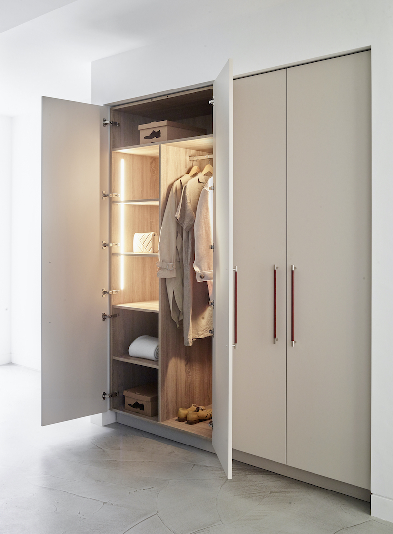 Custom fitted wardrobe with stone grey lacquered doors and copper bar handles revealing oak interior with LED shelf lighting, hanging rail, and organised storage compartments on grey stone flooring