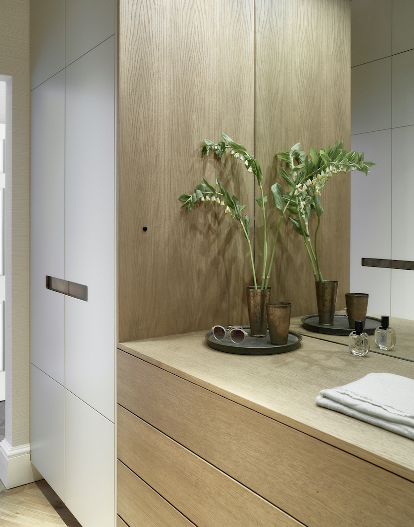 Custom fitted wardrobe with stone grey lacquered doors and copper bar handles revealing oak interior with LED shelf lighting, hanging rail, and organised storage compartments on grey stone flooring