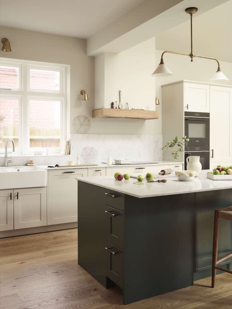 Contemporary classic kitchen with cream Shaker cabinets, brass lighting, marble worktops, farmhouse sink, and a deep-green island on warm wood flooring, styled with apples and simple accessories.