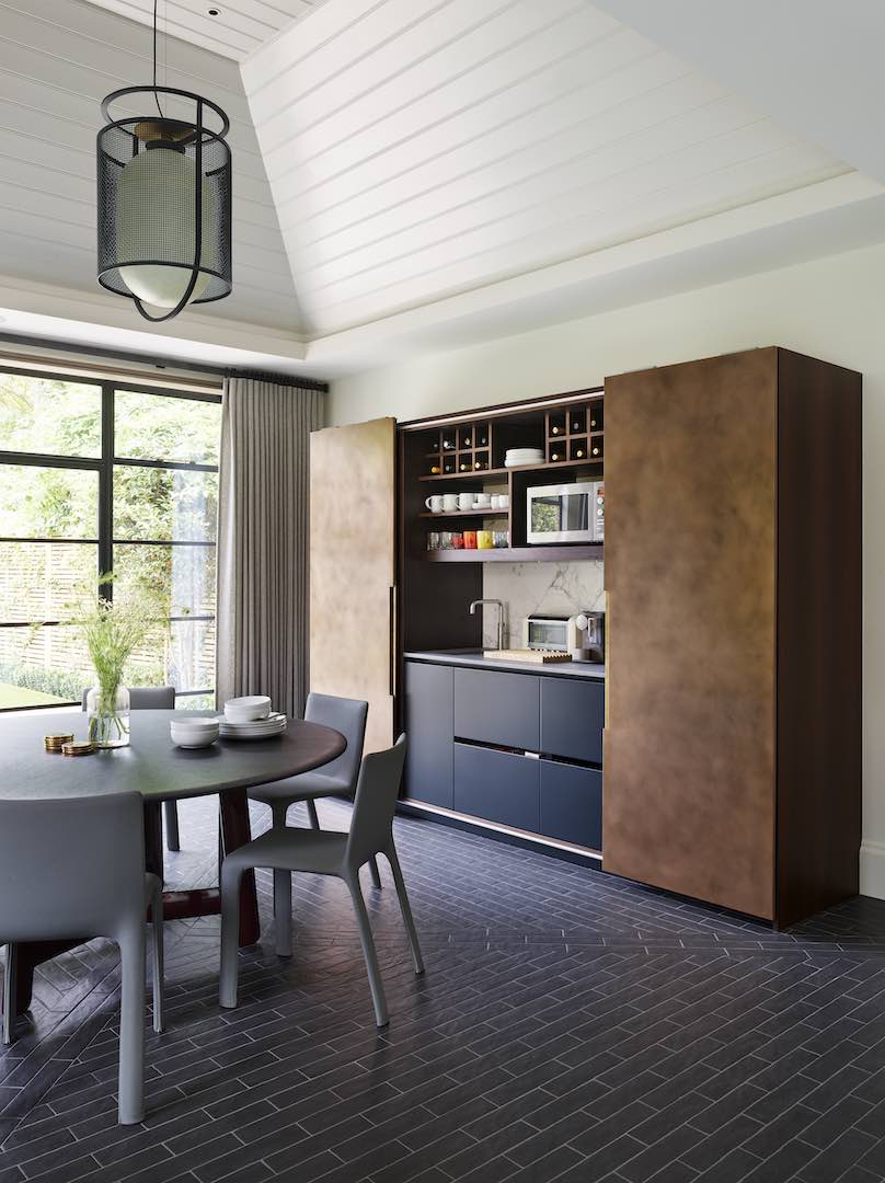 Pocket-door pantry wall in a dining area: patinated bronze doors open to a compact bar/kitchenette with navy drawers, sink, wine racks, and marble splashback beside a round table and Crittall-style windows.