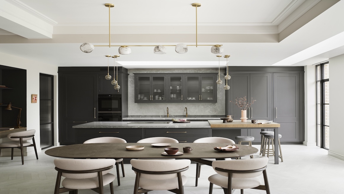 Modern grey kitchen with a large central island, integrated appliances, glass-fronted wall cabinets, and a dining area with upholstered chairs. Brass pendant lighting adds warmth and elegance to the space.