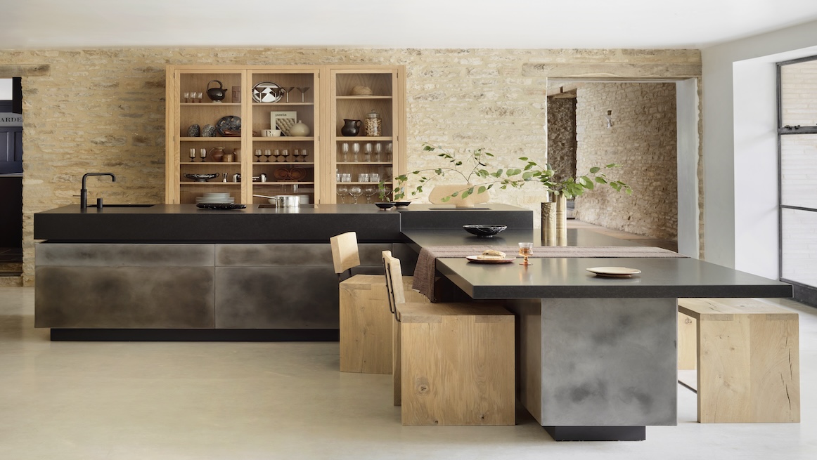 Rustic-modern space featuring a deep black stone island clad in patinated metal, an attached timber dining bar with block stools, pale stone walls, and floor-to-ceiling oak display cabinets filled with pottery and glassware.