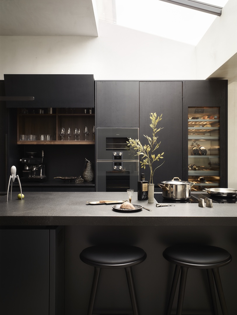 Contemporary dark kitchen with matt black cabinetry, integrated appliances, wine fridge, and open shelving. The central island in textured black stone includes an induction hob, bar seating, and modern accessories under a skylight.