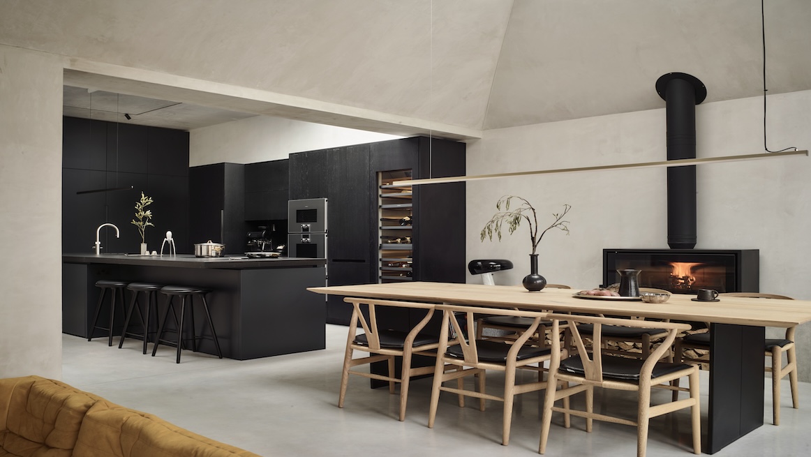 Minimalist black kitchen with a sleek island, integrated wine storage, and a wood-burning stove. A long wooden dining table with Scandinavian-style chairs sits in front, creating a striking contrast.