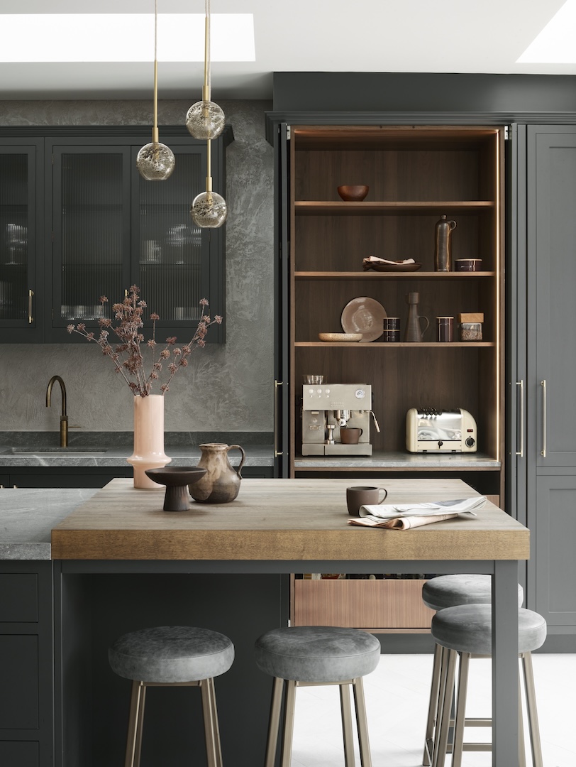 Charcoal grey kitchen with brass hardware, a smoked-glass pendant trio, a wood-topped breakfast bar and velvet stools, plus an open larder cabinet lined in walnut housing an espresso machine and ceramics.
