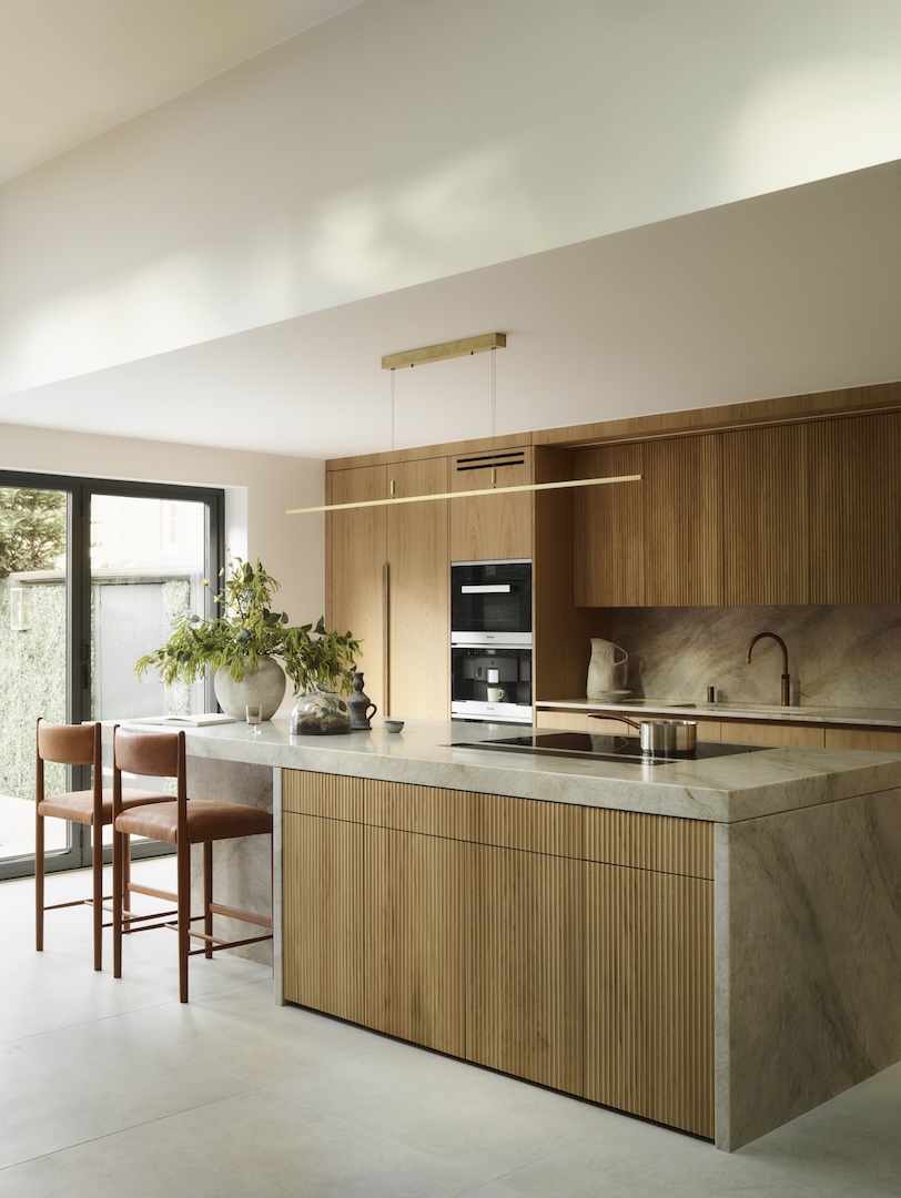 Contemporary kitchen with vertical wood panel cabinetry, a marble island, and built-in appliances. Natural light floods through full-height windows, highlighting the warm wood tones and stone surfaces.