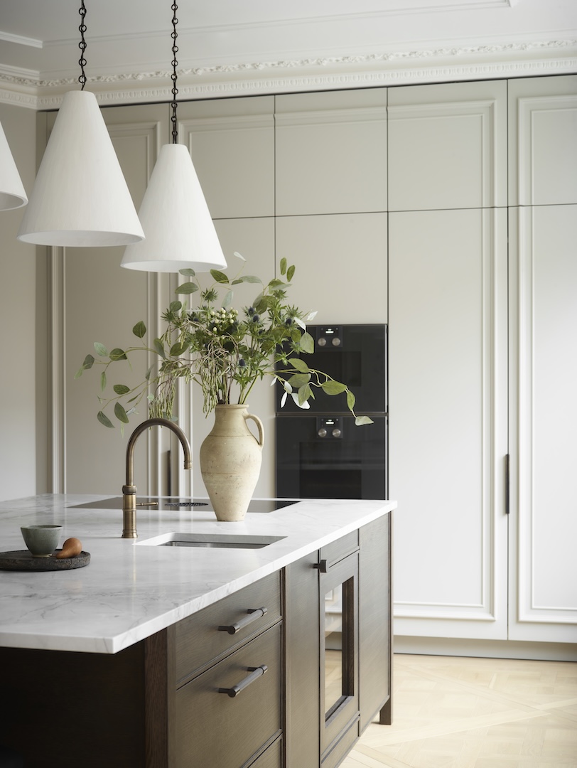 Elegant neutral kitchen showing tall cream paneled cupboards, a dark-stained island base topped with white marble, an old-brass faucet, three oversized white cone pendants, and a ceramic urn of greenery.