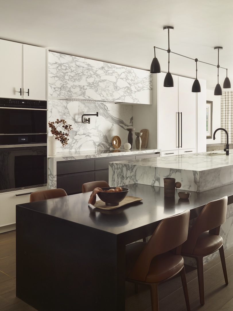 Luxury kitchen with dramatic white marble surfaces extending across cabinetry, splashback, and countertops. A dark dining table with leather chairs sits in the foreground beneath modern black pendant lights.