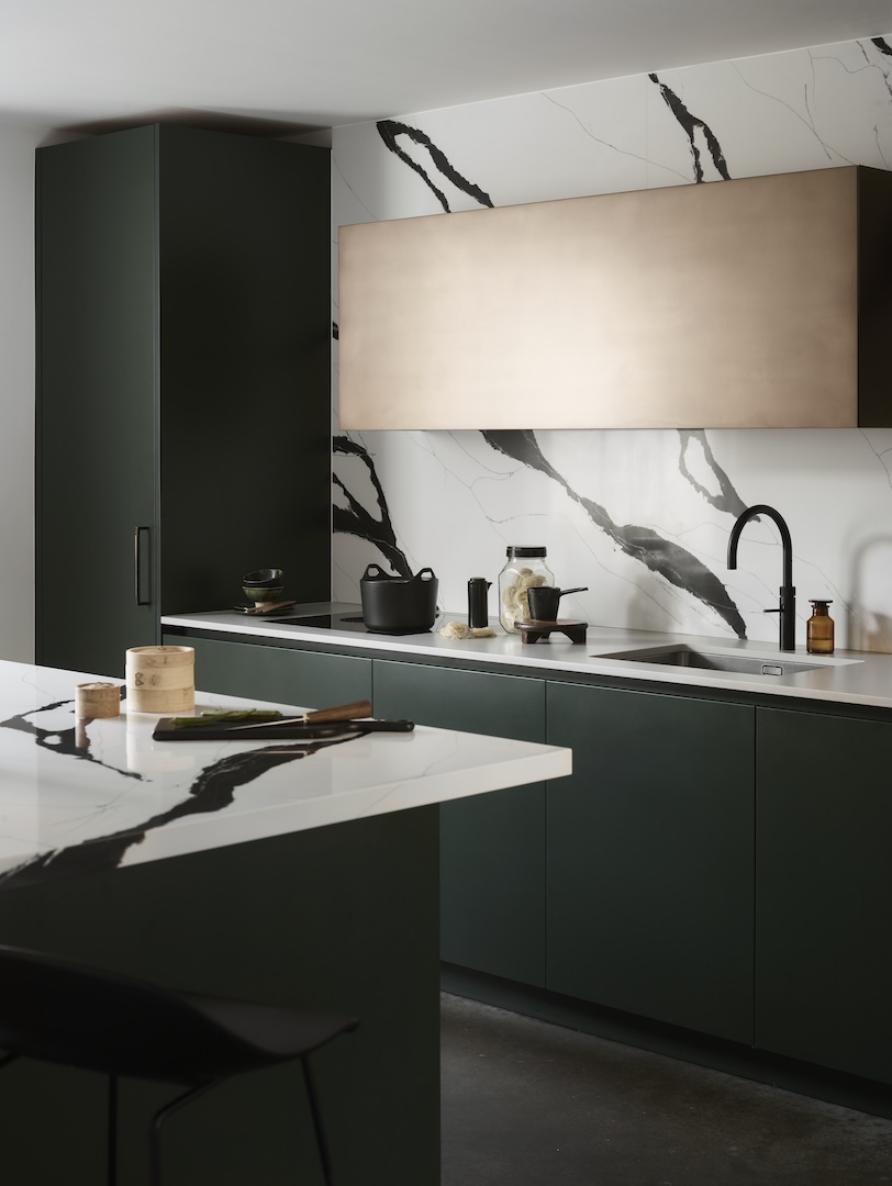 Sleek design of matte deep-green cabinets contrasted by a dramatic white quartz surface and backsplash streaked with bold black veining, capped with a large brushed-bronze hood panel and accented by black fixtures.