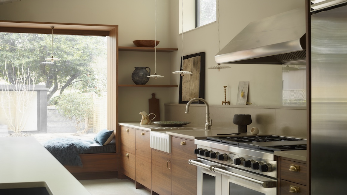 Warm, mid-century-inspired kitchen with walnut cabinetry, brass hardware, and open shelving. A stainless steel range cooker sits below a metal hood, with a window seat and garden views adding natural charm.