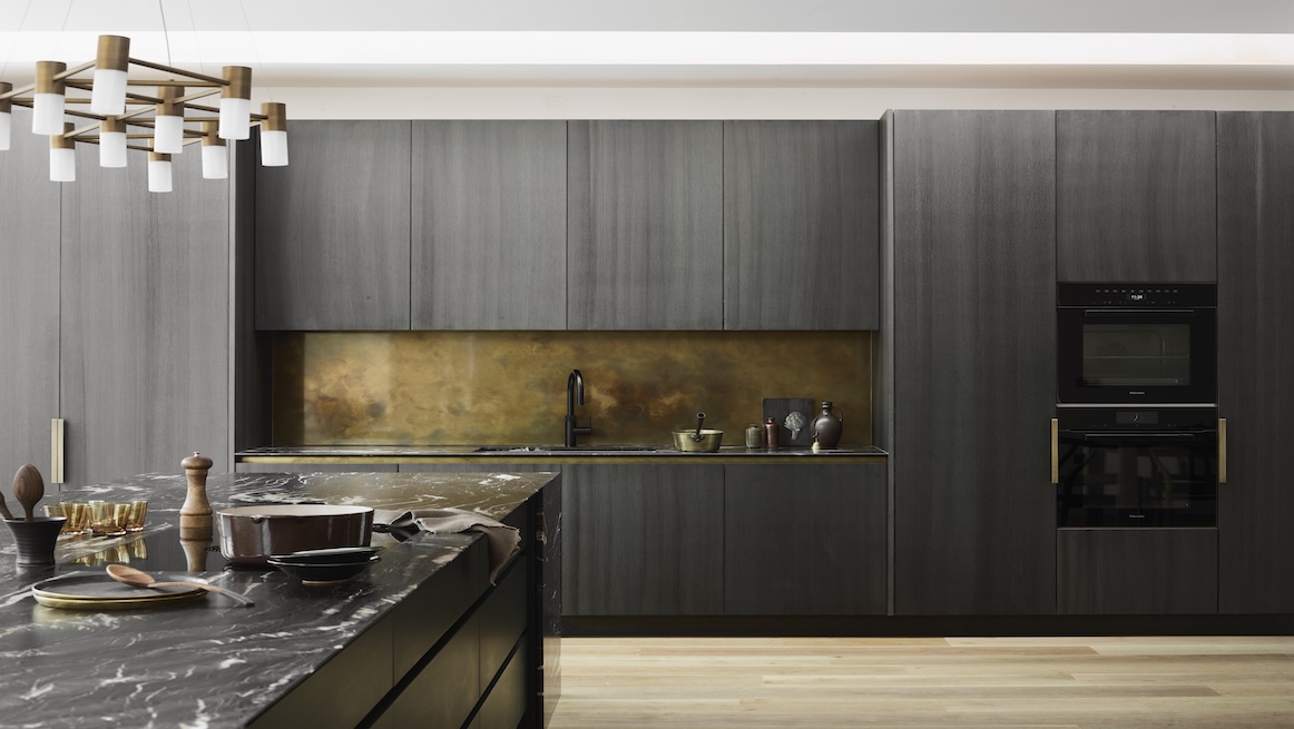 Moody kitchen clad in vertically grained dark-grey wood, a black marble island with vivid white veining, antiqued brass backsplash recess, integrated black appliances, and a brass-and-wood linear chandelier over light oak floors.