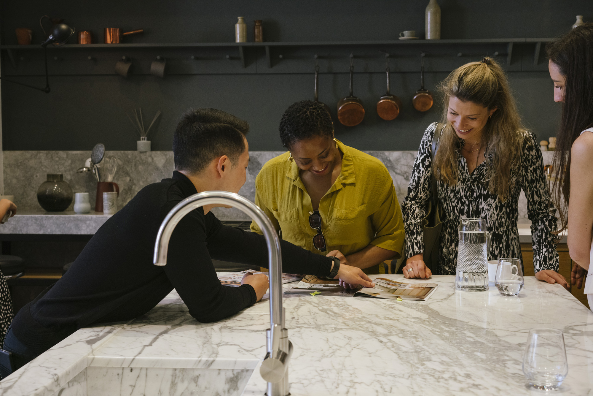 Professional kitchen consultation featuring "Trusted by Professionals" messaging with diverse group reviewing material samples at white marble island, copper pans displayed on dark wall, and "WORK WITH US" call-to-action button