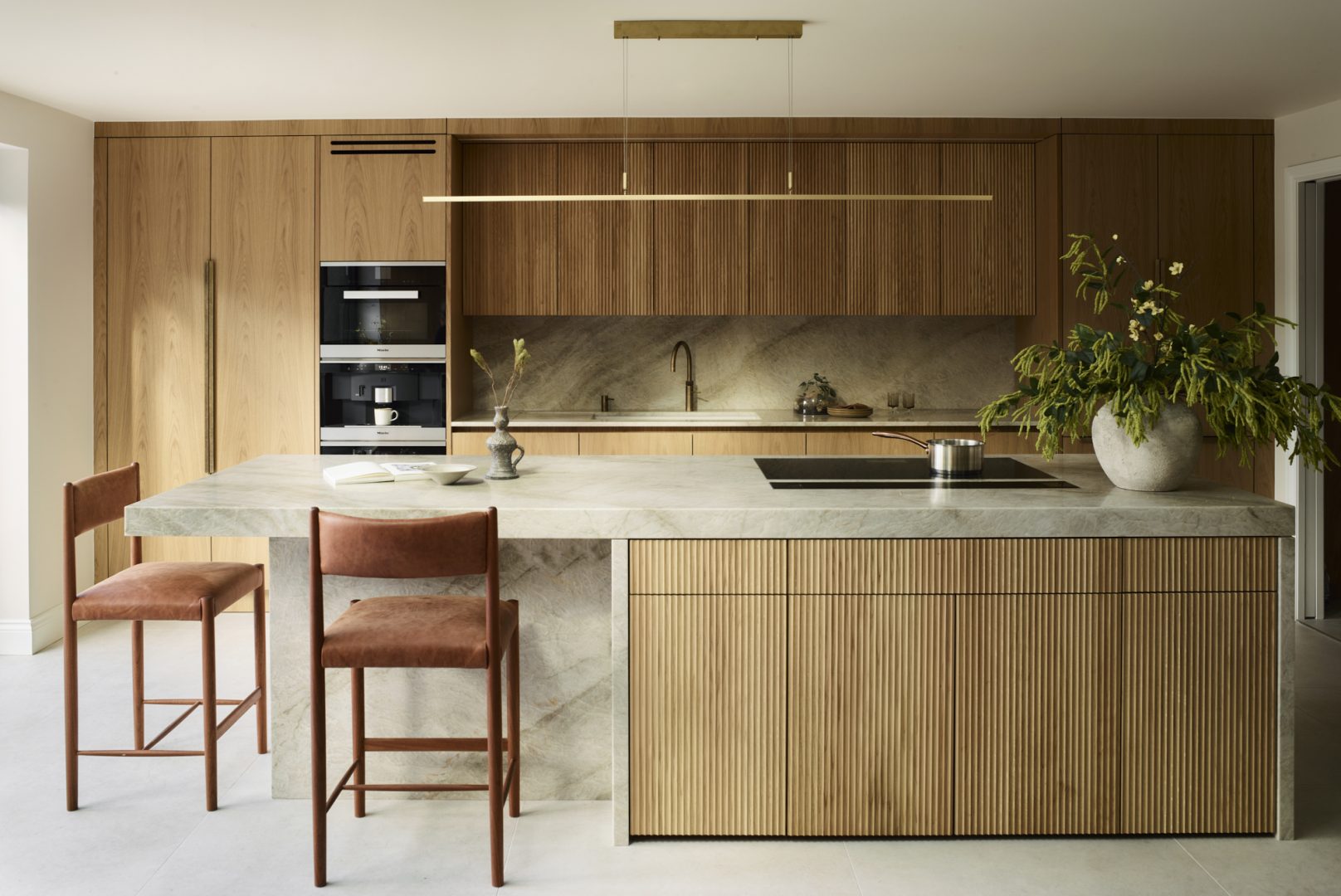 A modern kitchen featuring fluted oak cabinetry, a stone island with integrated hob, two leather bar stools, and a large planter with greenery, under a sleek brass pendant light.