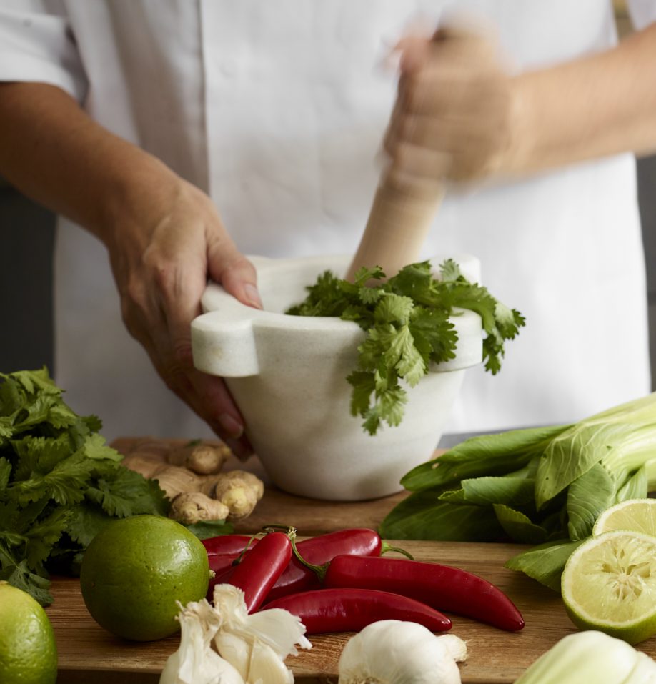 Hands using white marble mortar and pestle to prepare fresh herbs, surrounded by vibrant cooking ingredients including red chillies, limes, ginger, garlic, and leafy greens on timber worktop demonstrating culinary preparation in luxury kitchen setting