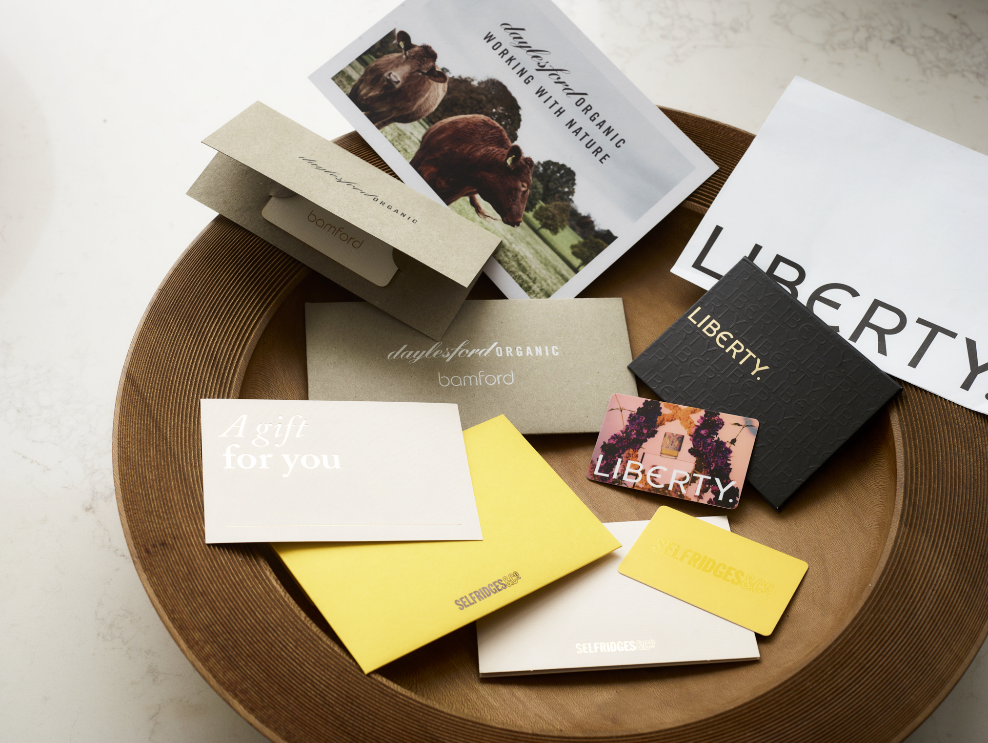 Marketing materials and stationery arranged on circular timber surface, including "Daylesford Organic" branded items, "LIBERTY" shopping bags, yellow envelopes, and various branded cards creating lifestyle brand collection display