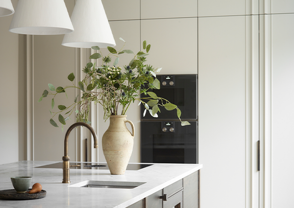 Contemporary white kitchen featuring brass tap, undermounted sink, white marble worktop, cream ceramic jug with fresh greenery, white cone pendant lights, and black built-in double oven creating serene minimalist cooking space