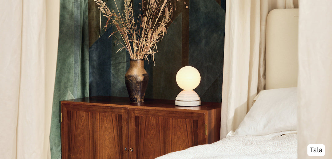 Bedroom corner with rich walnut storage cabinet featuring vertical grain detailing, ceramic vase with dried pampas grass, Tala spherical table lamp against emerald green textile backdrop