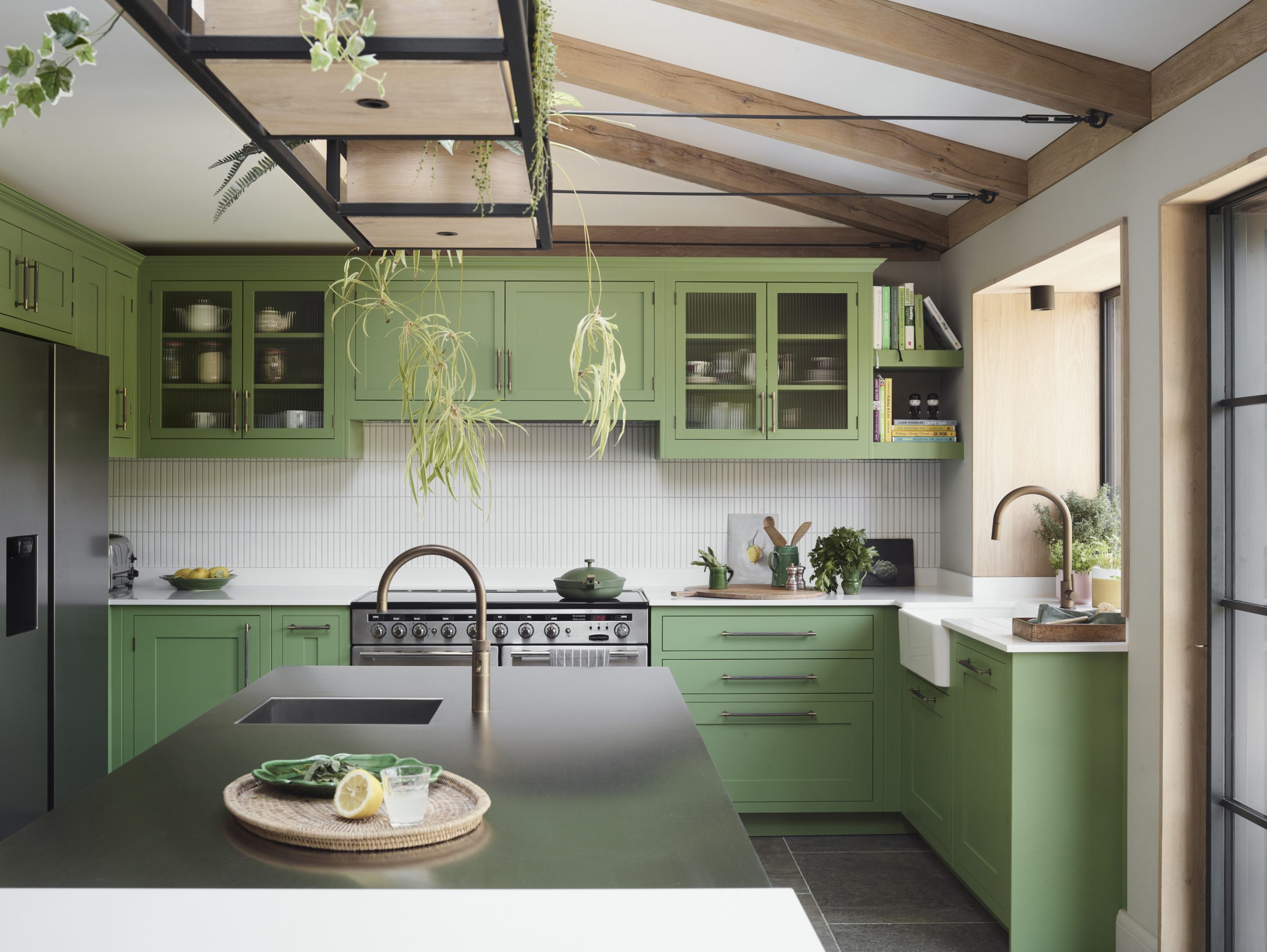 Bespoke sage green kitchen with exposed oak ceiling beams, featuring shaker-style cabinetry, white vertical tile splashback, and charcoal island with brass bridge tap, suspended metal shelving displaying trailing plants, slate flooring