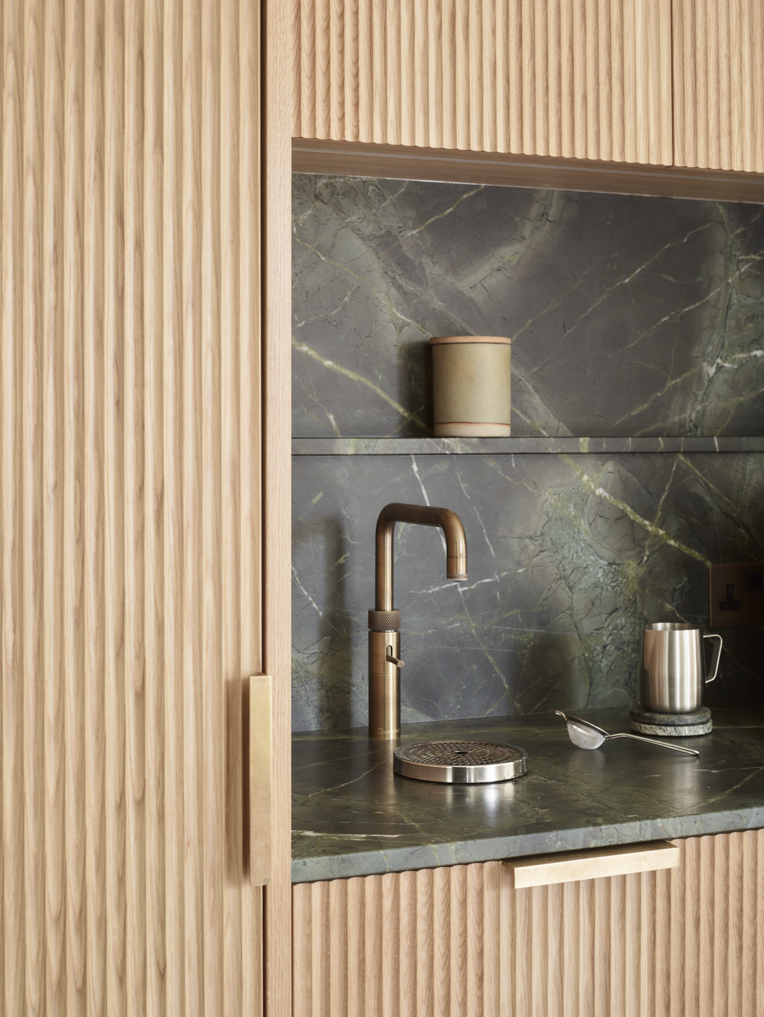 Luxury kitchen detail showing fluted timber cabinetry with Verde Guatemala marble worktop and splashback, antique brass square-profile tap, bespoke oak storage niche with integrated shelf, demonstrating premium material harmony