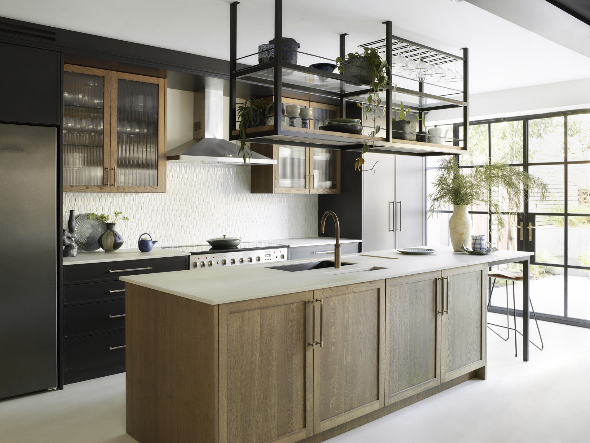 Modern kitchen combining charcoal shaker cabinetry with natural oak island, white geometric tile splashback, suspended black metal shelving system, floor-to-ceiling Crittall-style windows, integrated appliances and brass hardware