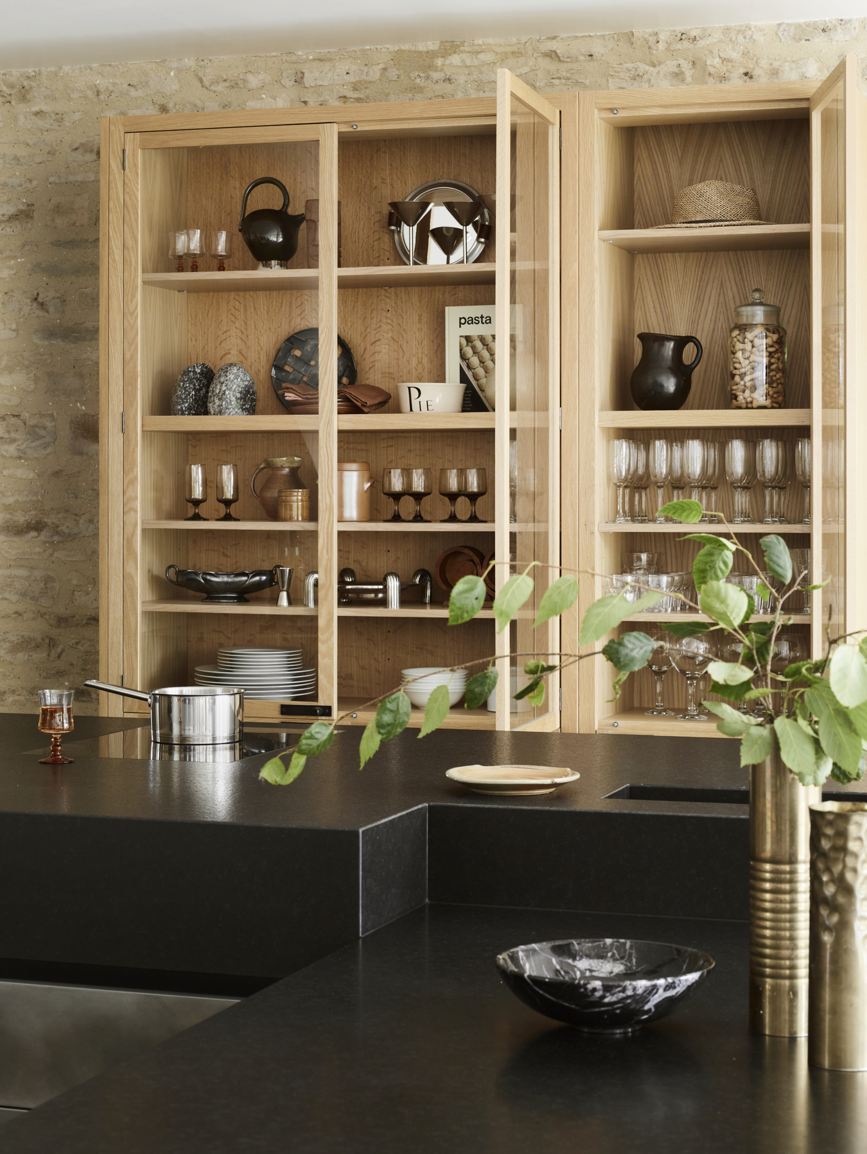 Bespoke oak display cabinetry with glazed doors revealing organised dishware and glassware, exposed London brick wall, black granite worktop with brass tap, demonstrating clever storage solutions and material contrasts
