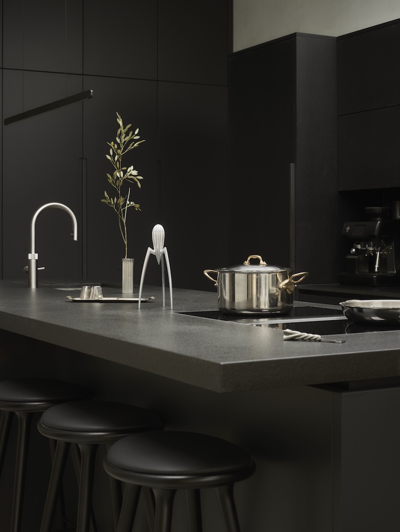 Monochromatic black kitchen island with integrated induction hob, chrome tap, bar seating, and built-in coffee machine, exemplifying sophisticated minimalist design