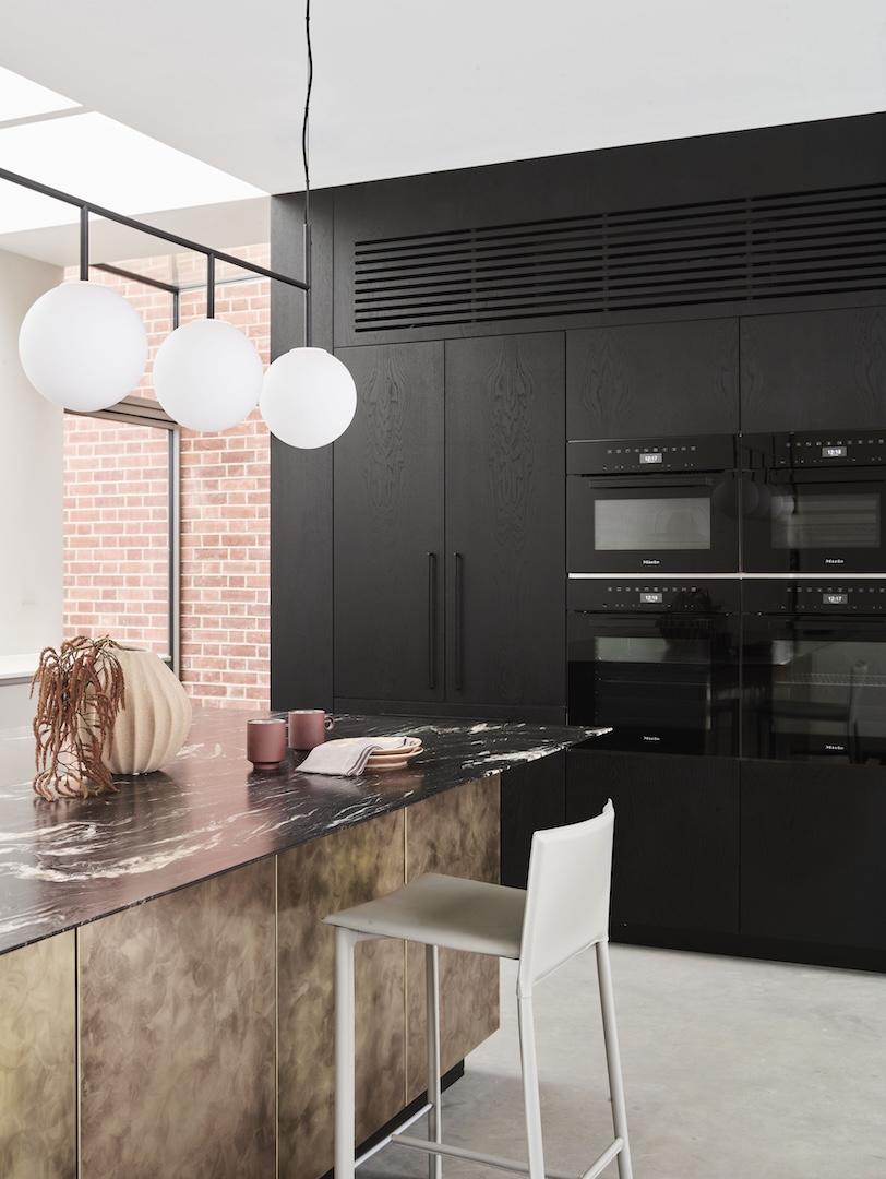 Two-tone bespoke kitchen combining ebonised oak tall cabinets with integrated Miele ovens, weathered brass island base, dramatic Nero Marquina marble worktop, and globe pendant lighting