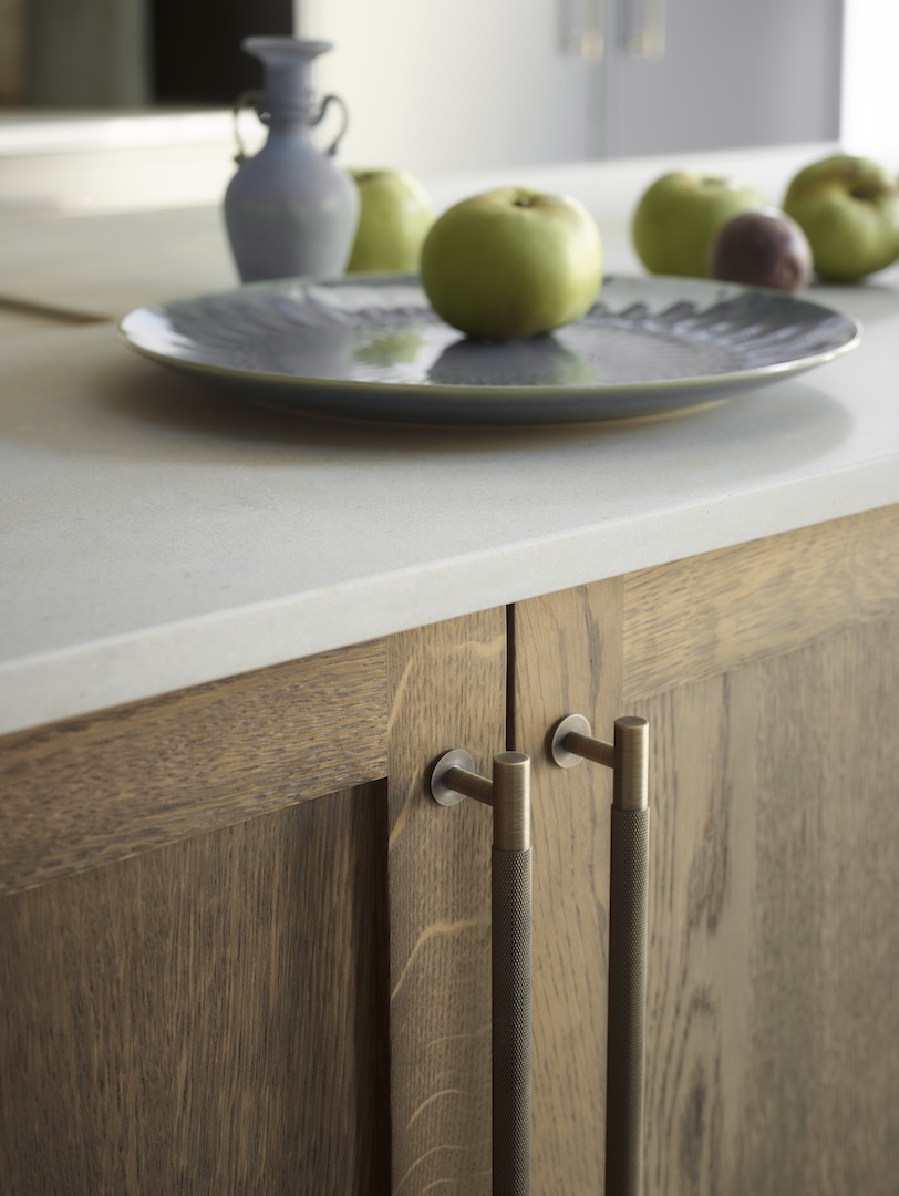 Natural oak kitchen cabinetry detail with pristine white composite worktop and bronze knurled handles, styled with grey ceramic platter and green apples