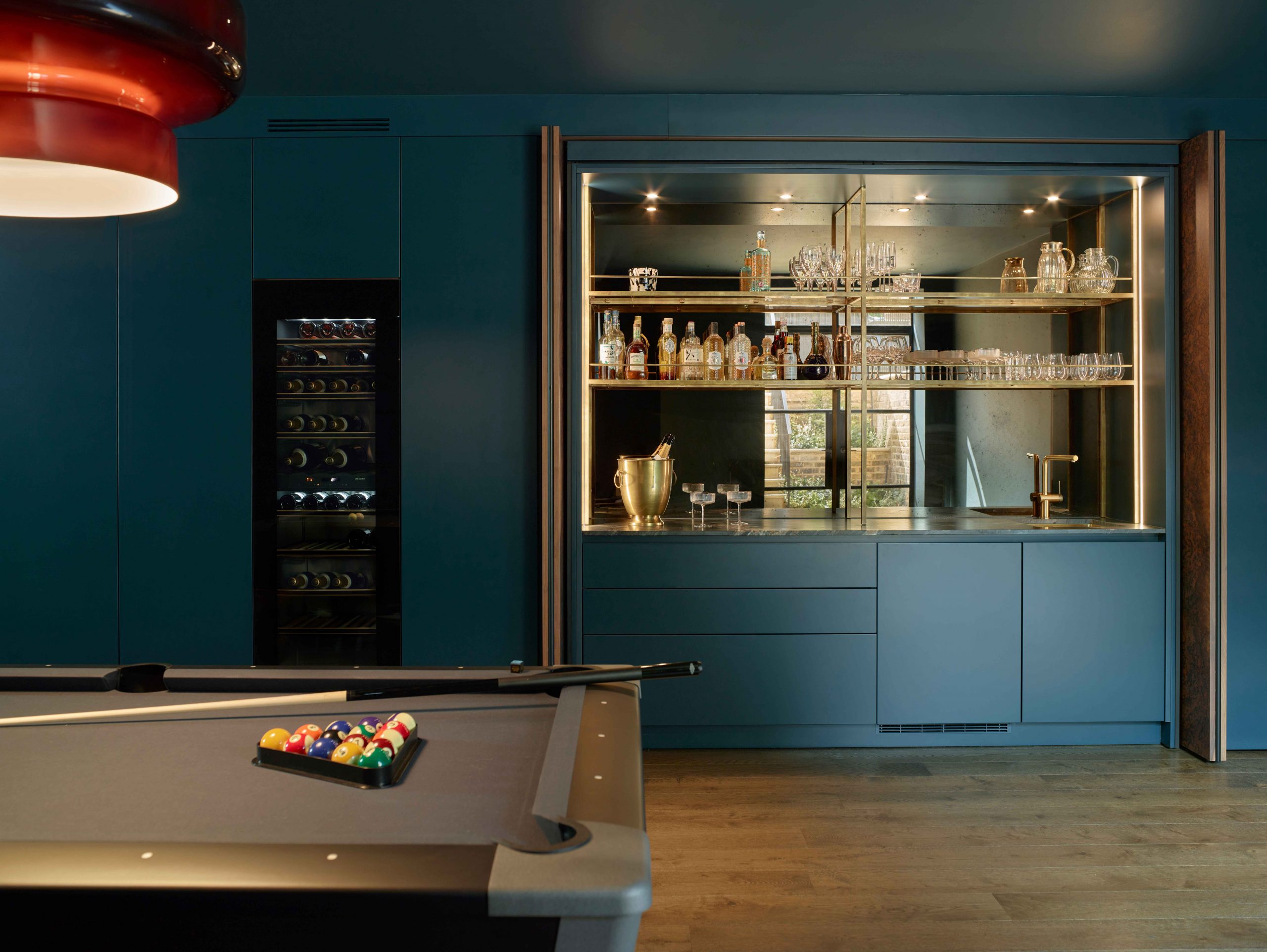 Bespoke entertainment room with deep teal cabinetry featuring illuminated cocktail bar with brass-framed glass shelving, mirrored splashback displaying crystal glassware and spirits, integrated wine cooler, contemporary pool table with herringbone oak flooring