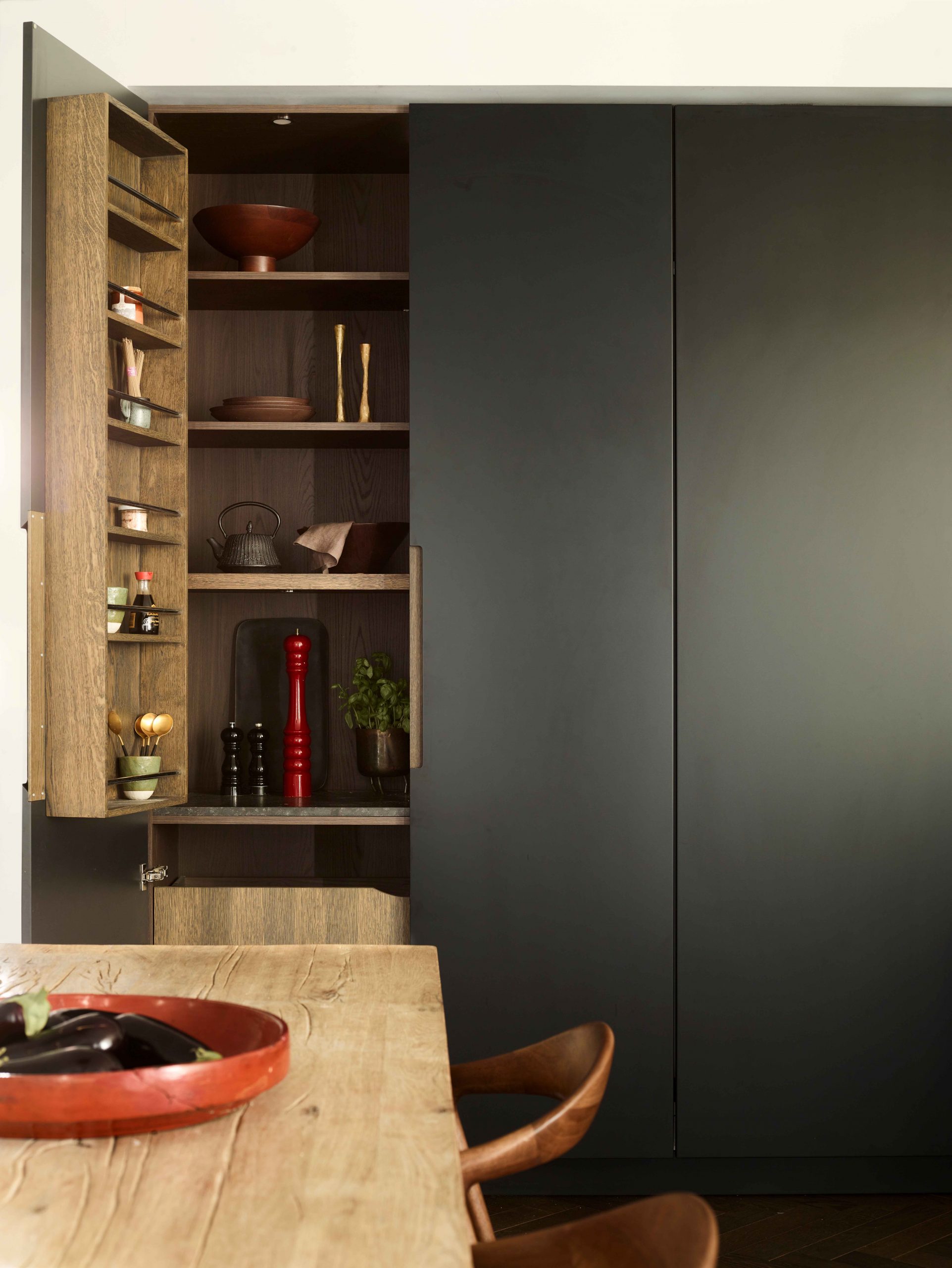 Bespoke kitchen storage with charcoal grey pocket doors opening to reveal oak-lined pantry shelving displaying ceramics, cast iron teapot, spices and condiments, beside natural timber dining table with tan leather chairs
