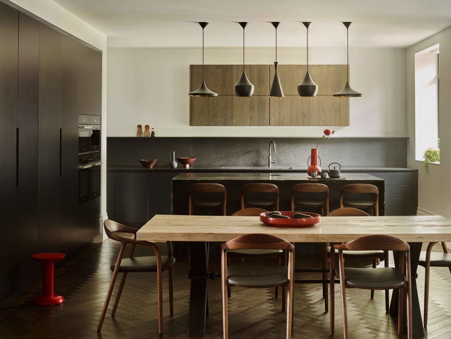 Contemporary bespoke kitchen with charcoal grey cabinetry, black granite worktop and splashback, natural fluted timber wall units, five sculptural black pendant lights above island with leather bar stools, oak dining table on herringbone parquet flooring