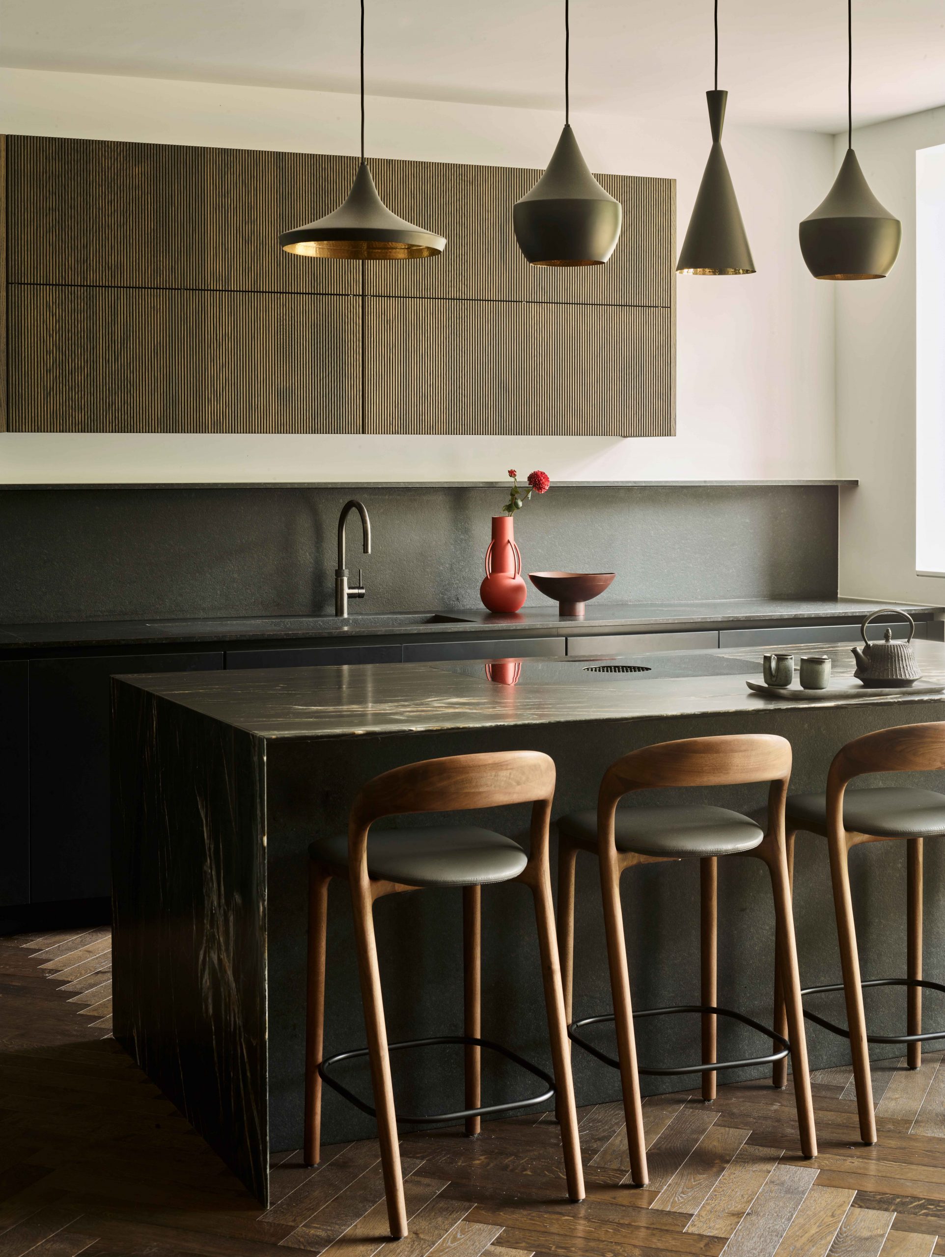 Bespoke kitchen island with black granite worktop, four sculptural matte black pendant lights with brass interiors, natural timber bar stools with grey upholstery, fluted oak wall units above grey stone splashback, herringbone parquet flooring