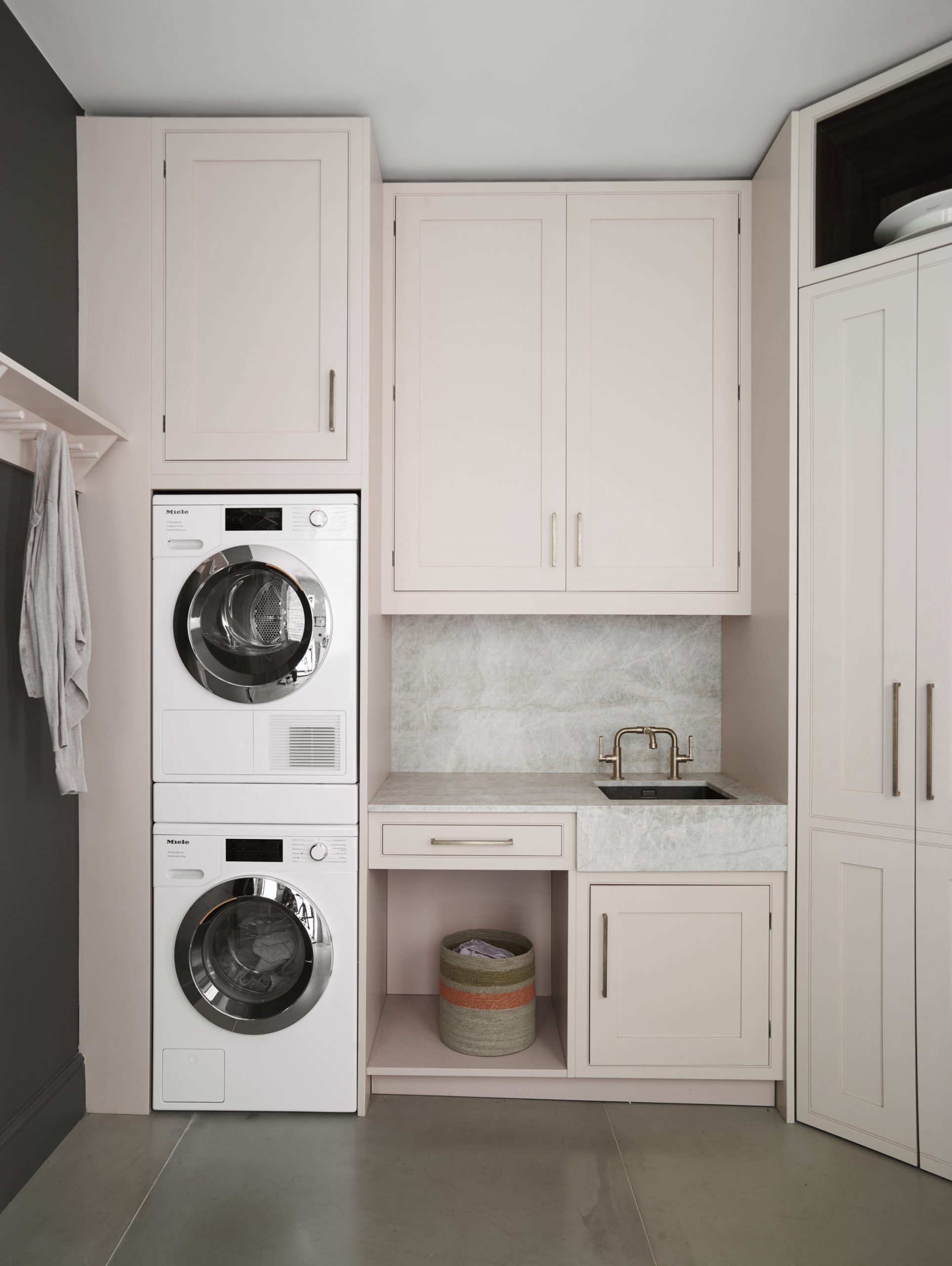 Luxury blush pink shaker-style utility room with stacked Miele washer and dryer, Carrara marble worktops and splashback, brass bridge tap, bespoke cabinetry with champagne bronze handles, and built-in open shelving