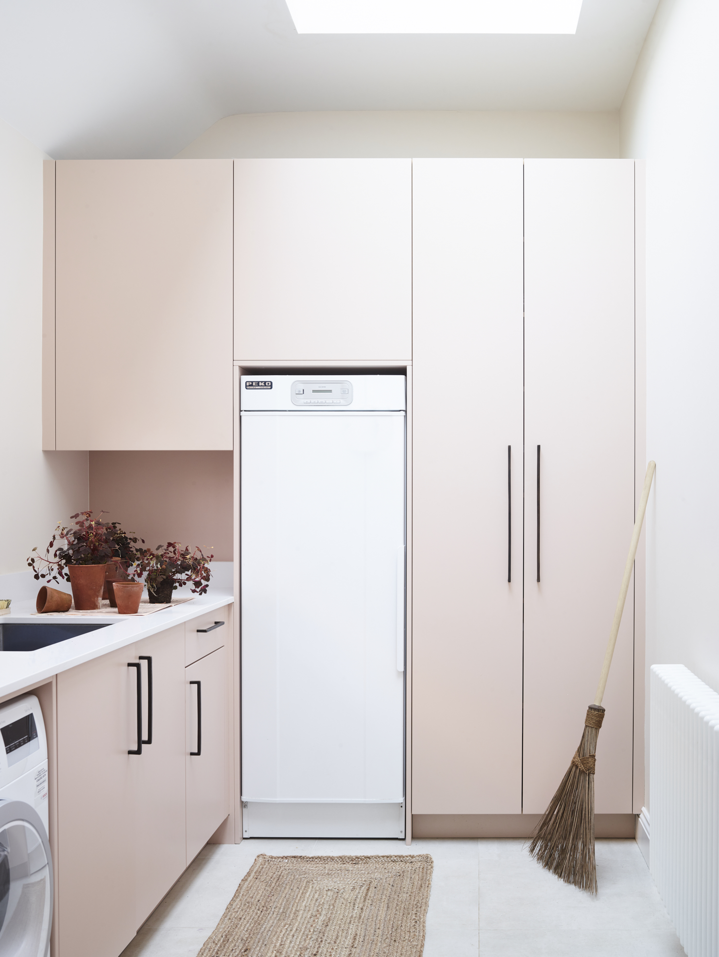 Light-filled pink utility room beneath skylights with integrated tall freezer, shaker-style cabinetry, white composite worktops, terracotta planters, natural jute rug, and seamless handleless storage solutions