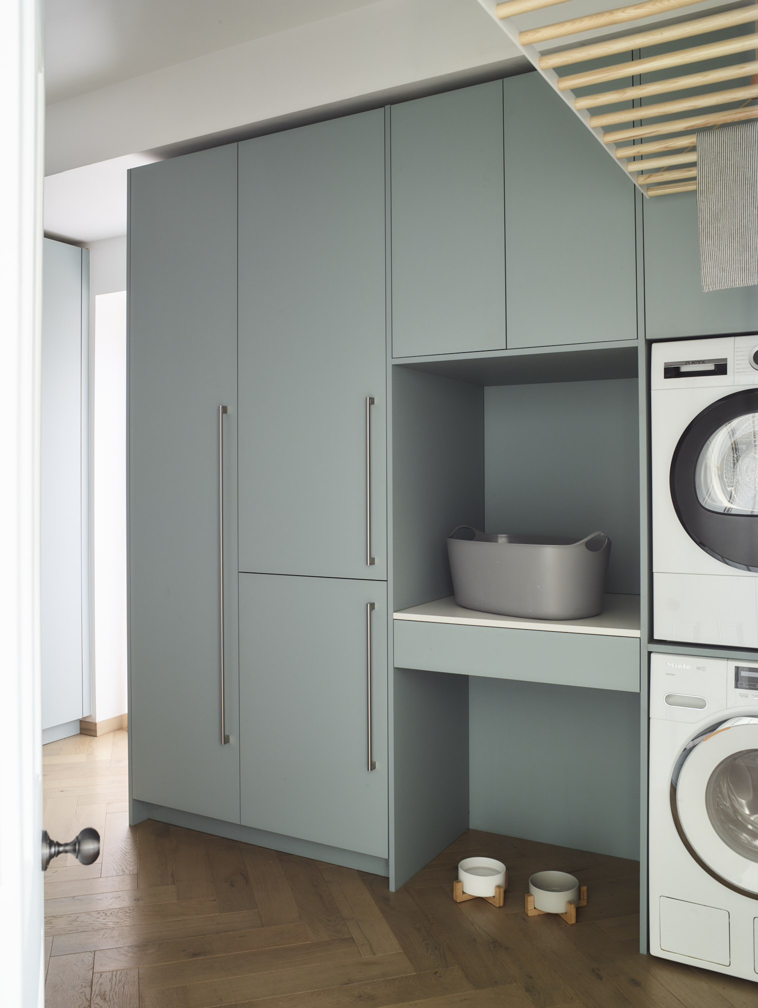 Duck egg blue utility room with stacked washing machines, floor-to-ceiling bespoke storage, open shelving with grey storage baskets, exposed timber ceiling beams, herringbone oak flooring, and integrated pet feeding station