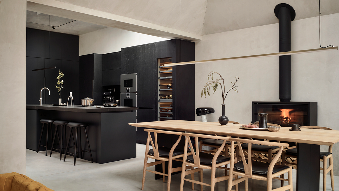 Bespoke charcoal black kitchen with handleless cabinetry, breakfast bar island, and natural oak dining table alongside contemporary fireplace in open-plan living space