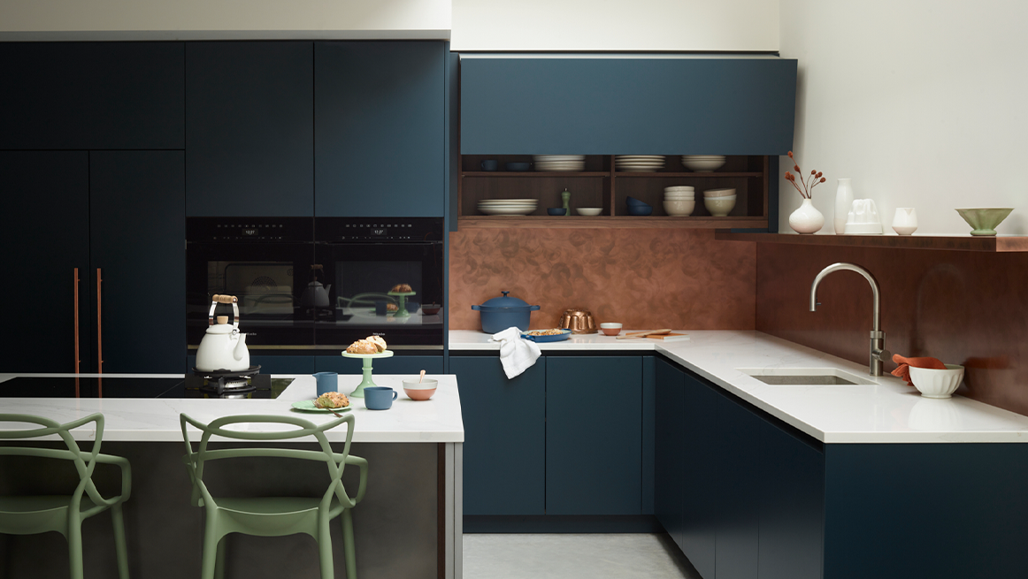Navy blue L-shaped kitchen with terracotta splashback, white composite worktops, integrated appliances, and breakfast bar with sage green contemporary dining chairs