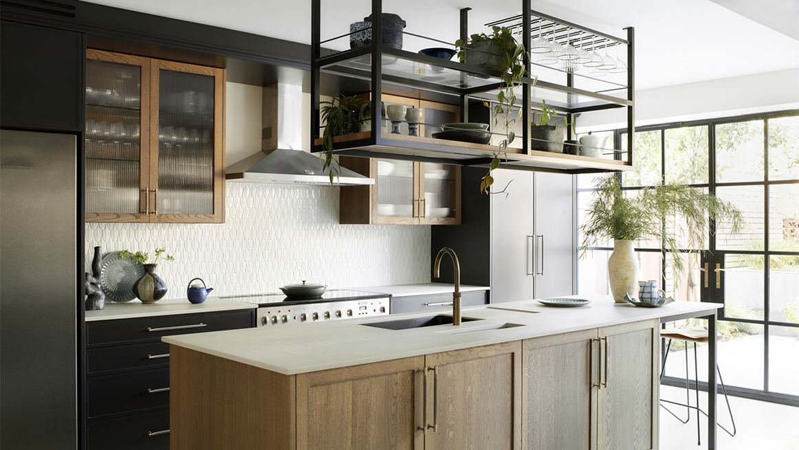 Contemporary kitchen combining black base units, natural oak island with integrated hob, suspended metal shelving system, white textured splashback tiles, and floor-to-ceiling Crittall-style windows