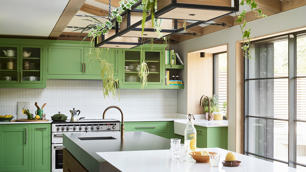 Vibrant green shaker kitchen with suspended black metal frame ceiling planter, professional range cooker, white composite worktops, copper bridge tap, ribbed glass wall cabinets, and floor-to-ceiling glazing