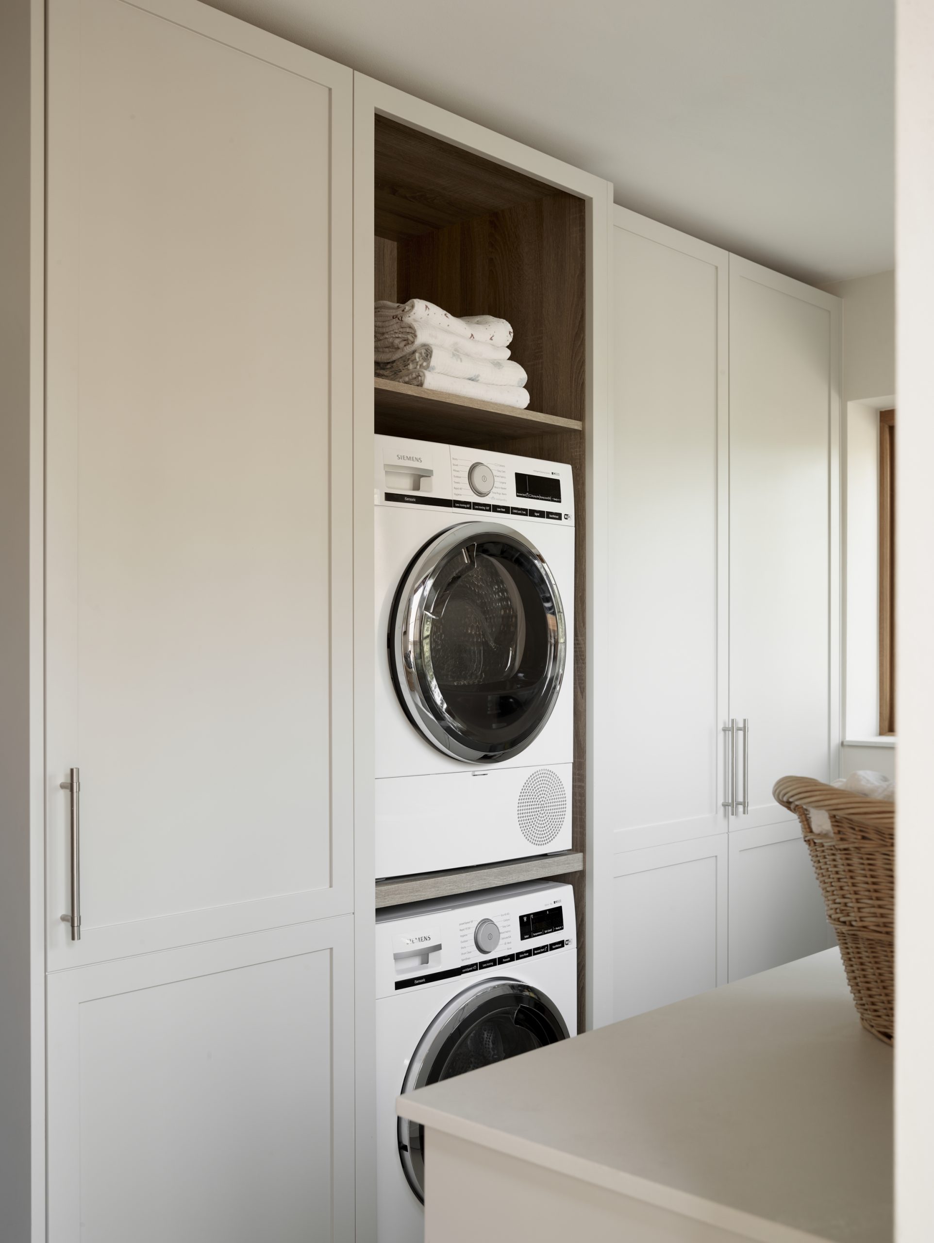 Cream shaker-style utility room with floor-to-ceiling bespoke cabinetry, integrated stacked Siemens washer and dryer, walnut timber shelving alcove with folded white towels, brushed steel handles, pull-out ironing board, and woven laundry basket