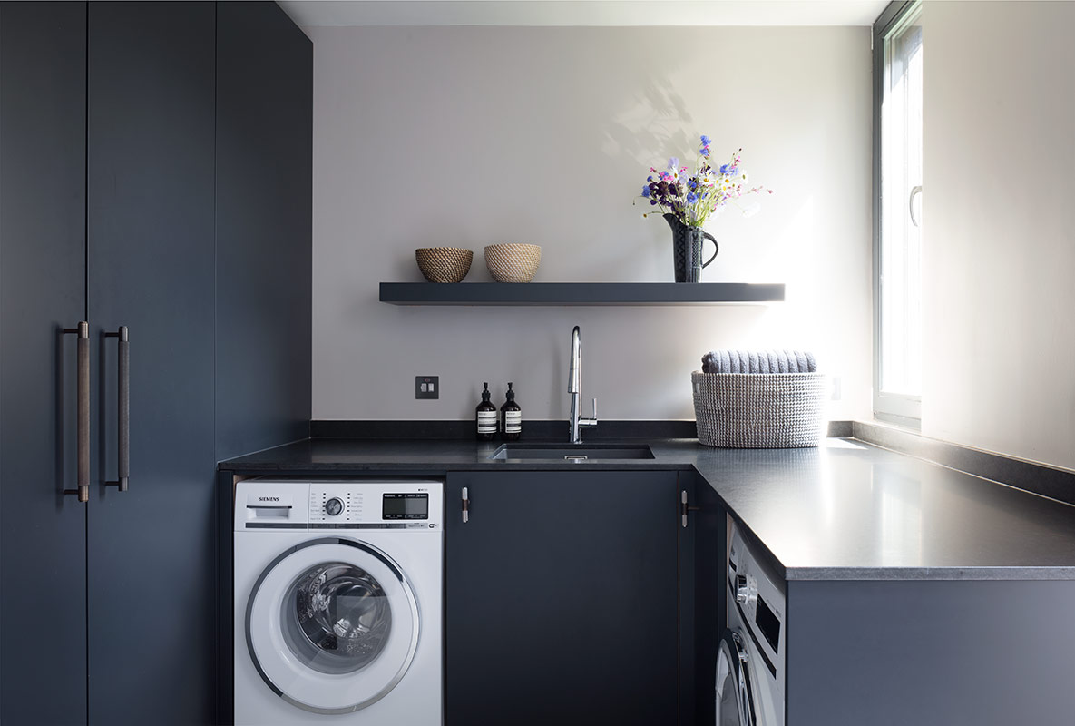 Bespoke charcoal grey utility room featuring handleless cabinetry with integrated washing machine, black granite worktop, floating shelf styled with ceramic bowls and purple wildflowers, chrome mixer tap, and floor-to-ceiling storage