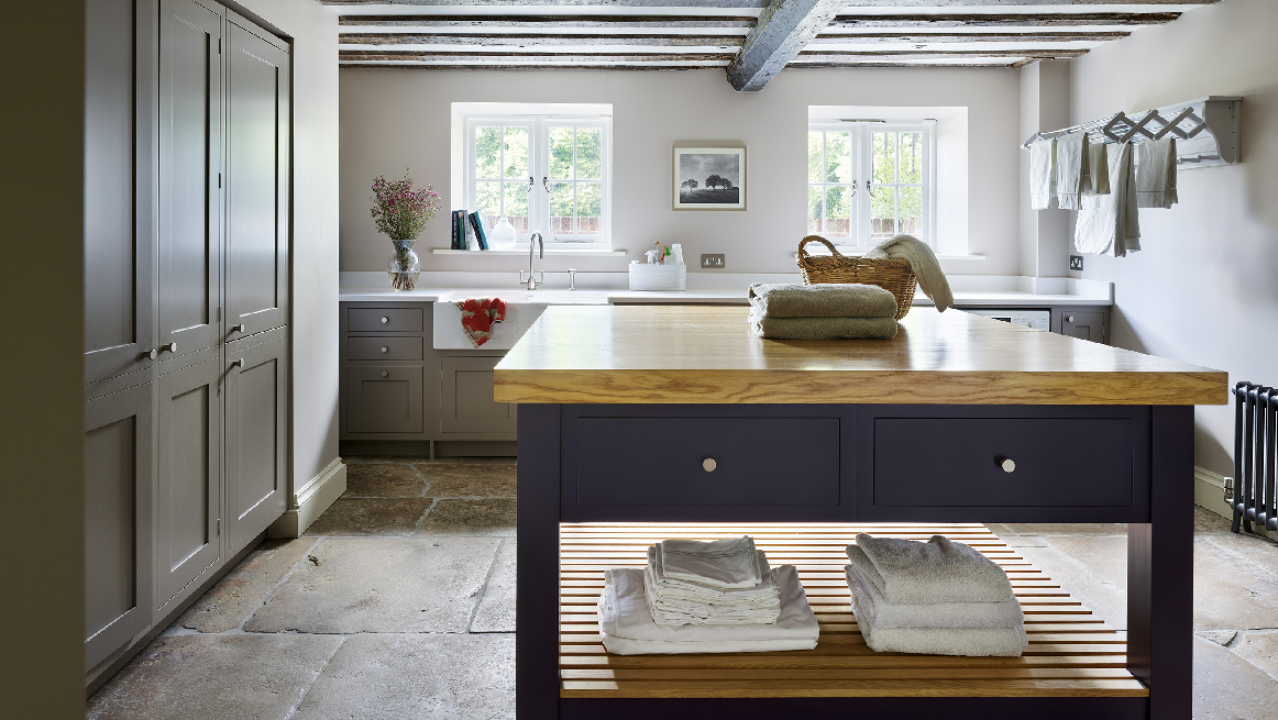 Bespoke country utility room with grey shaker cabinetry, navy blue island featuring solid oak worktop and open linen storage, exposed whitewashed ceiling beams, and aged limestone flooring in a period property
