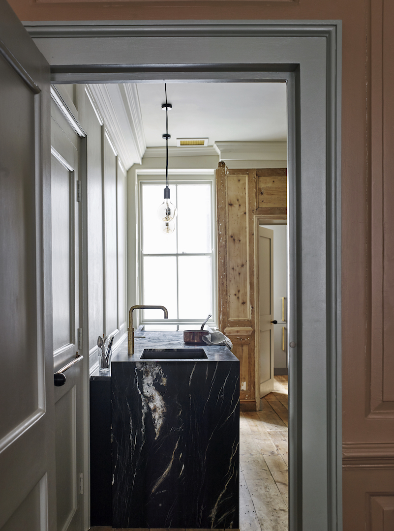 Black marble kitchen island with brushed brass tap framed by grey painted doorway leading to bright natural timber joinery in period property restoration