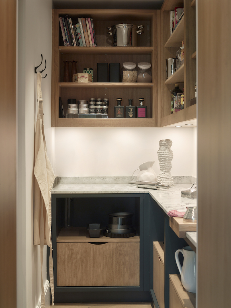 Compact butler's pantry with natural oak open shelving displaying cookbooks and storage jars, white marble worktop, deep blue base units with oak drawer boxes, undermount LED lighting, and wall-mounted coat hooks