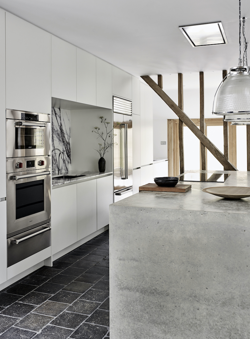 Contemporary white handleless kitchen with polished concrete island worktop, exposed oak structural beams, and integrated professional appliances in converted barn
