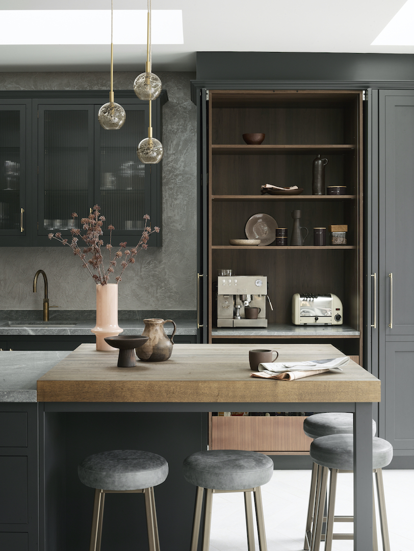 Sophisticated charcoal grey kitchen with reeded glass cabinet doors, polished concrete splashback, thick oak breakfast bar, concealed coffee station with espresso machine and toaster, grey velvet bar stools with brass legs, and bubble glass pendant lights