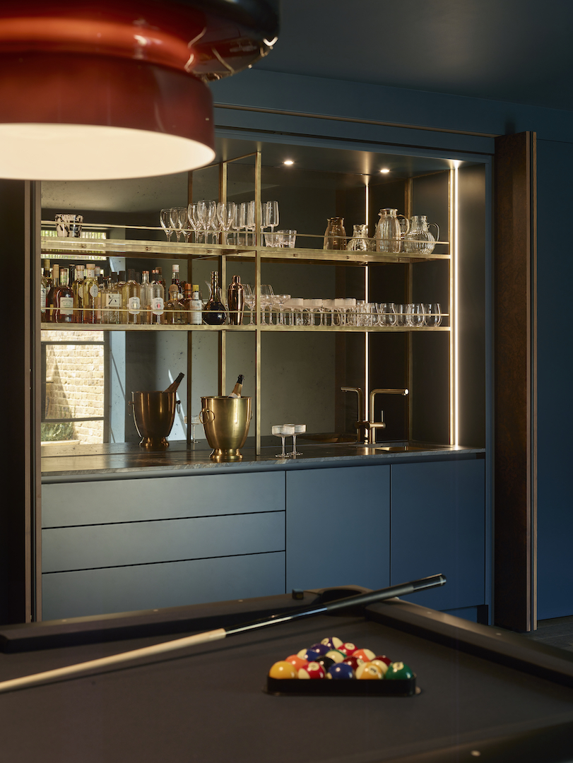 Bespoke home bar in deep blue with brass-framed glass shelving displaying spirits and glassware collection, integrated sink with brass tap, champagne buckets, LED strip lighting, dark walls, and snooker table in foreground